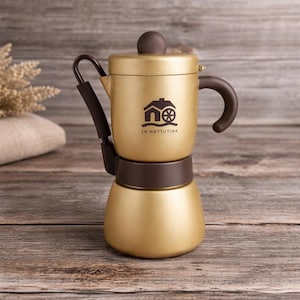 May include: A gold-colored stovetop espresso maker with brown accents. The coffee maker has a rounded base and a cylindrical upper chamber. The handle and knob are brown. The logo "LA MATTUTINA" is printed on the front.