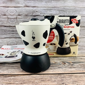 Könnte beinhalten: Ein Bialetti Mukka Express Herd-Cappuccinobereiter mit weißem Gehäuse und schwarzen Kuhflecken, einem schwarzen Griff und einem schwarzen Boden. Das Produkt wird mit der Originalverpackung aus Karton und einer Bedienungsanleitung gezeigt.
