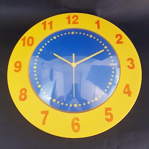 May include: A round wall clock with a yellow frame and a blue face. The clock has orange numbers and yellow hands. The clock face has a dotted circle around the edge. The brand name "bino" is printed on the clock face.