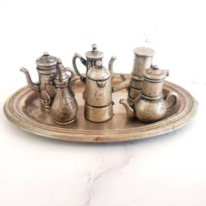 Può includere: Una collezione di piccole caffettiere argentate e un vassoio ovale. Le caffettiere variano nel design, con beccucci, manici e coperchi. Il vassoio ovale ha un aspetto leggermente appannato. La disposizione è su una superficie bianca.