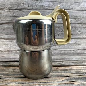 Könnte beinhalten: Ein Espressokocher aus Edelstahl mit polierter Oberfläche sowie einem Deckel und Griff aus cremefarbenem Kunststoff. An der Seite des Kessels befindet sich die eingravierte Aufschrift Lagostina Made in Italy. Das Design umfasst einen breiten Boden.