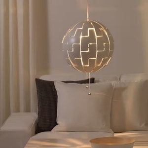 Pode incluir: Uma luminária pendente branca em forma de esfera com recortes. A luz está pendurada sobre um sofá branco com duas almofadas. A luz está acesa e projeta um brilho quente.