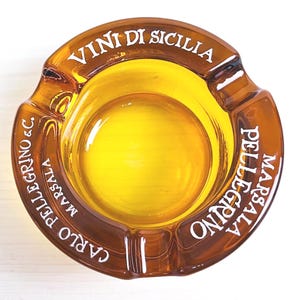 Peut inclure: Cendrier en verre ambré avec le texte "VINI DI SICILIA CARLO PELLEGRINO & C. MARSALA" et "MARSALA PELLEGRINO" sur les côtés.