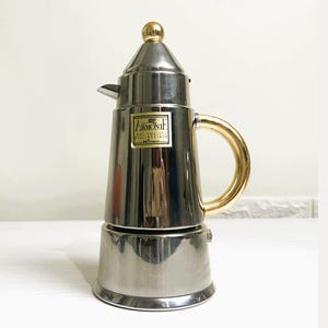 Pode incluir: Uma cafeteira de espresso de aço prateado e dourado com alça dourada e um rótulo que diz "ARMONIA" em letras douradas. O rótulo também diz "ITAL QUALITY" e "MADE IN ITALY".