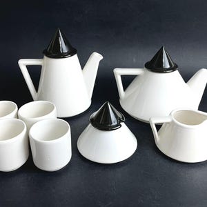 Può includere: Un servizio da tè composto da due teiere bianche con coperchi conici neri, quattro tazze cilindriche bianche, una zuccheriera bianca con coperchio conico nero e una lattiera bianca. Ogni pezzo presenta una finitura in ceramica liscia.