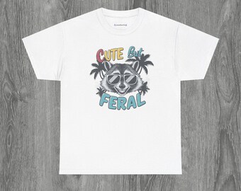 Op de afbeelding: Wit T-shirt met een afbeelding van een wasbeer met een zonnebril en palmbomen. De tekst "Cute but Feral" is onder de wasbeer gedrukt.