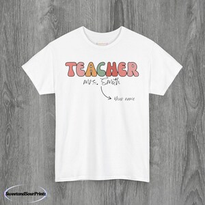 May include: White crew neck t-shirt featuring the word "TEACHER" in a retro font, with "Mrs. Smith" below. An arrow indicates a space for a name. The shirt is made of a soft material.