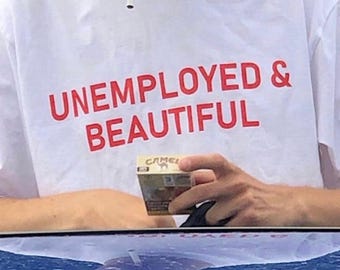 Könnte beinhalten: Ein weißes T-Shirt mit dem Text "UNEMPLOYED & BEAUTIFUL" in roten Buchstaben. Eine Person hält eine Schachtel Zigaretten.