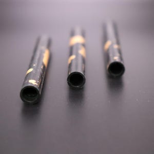 May include: Three black cylindrical objects with a hollow center and gold-colored accents. The objects are arranged horizontally against a dark background, with the openings facing the viewer. The objects appear to be small tubes.