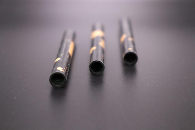 May include: Three black cylindrical objects with a hollow center and gold-colored accents. The objects are arranged horizontally against a dark background, with the openings facing the viewer. The objects appear to be small tubes.