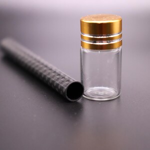 May include: A clear glass vial with a gold-colored cap and a black, textured tube. The vial is cylindrical and empty. The tube is black with a geometric pattern. The items are on a dark surface.