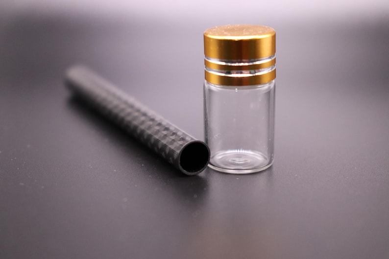 May include: A clear glass vial with a gold-colored cap and a black, textured tube. The vial is cylindrical and empty. The tube is black with a geometric pattern. The items are on a dark surface.