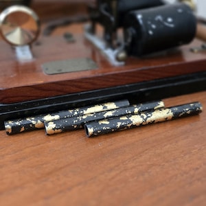 May include: Close-up of black tubes with gold leaf accents, likely for a vintage or antique device. The tubes are arranged on a wooden surface, with a blurred background featuring other mechanical components.