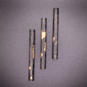 May include: Three dark, cylindrical objects with gold-colored accents are arranged vertically against a dark gray background. The objects appear to be made of a dark material, possibly metal or a composite, and have a smooth, reflective surface.