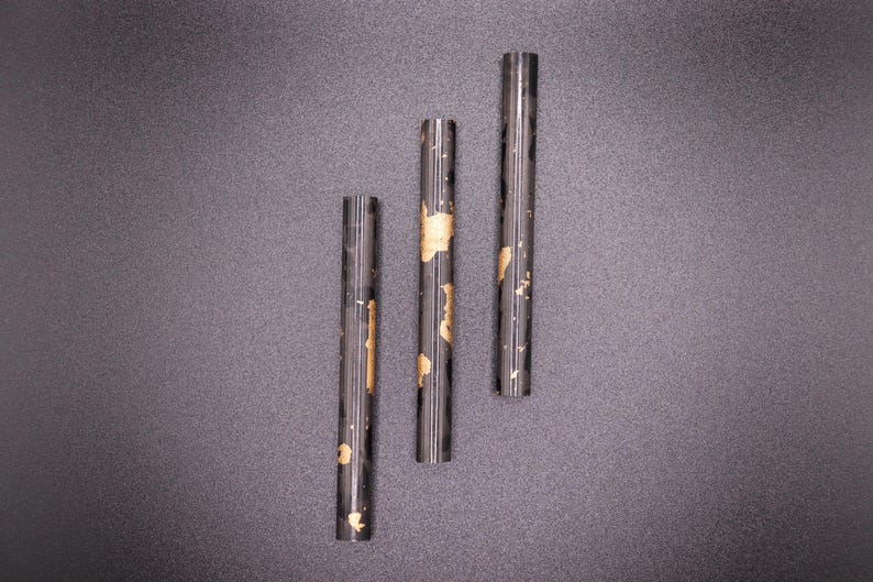 May include: Three dark, cylindrical objects with gold-colored accents are arranged vertically against a dark gray background. The objects appear to be made of a dark material, possibly metal or a composite, and have a smooth, reflective surface.