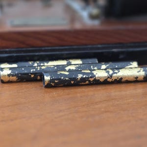 May include: Close-up of three black and gold decorative pen-like objects. The objects have a black base with gold leaf accents. The items are resting on a wooden surface, with a blurred wooden and black background.