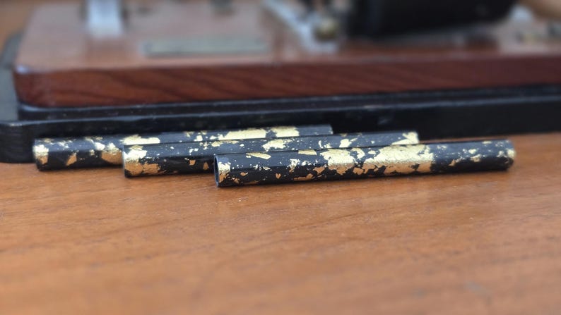 May include: Close-up of three black and gold decorative pen-like objects. The objects have a black base with gold leaf accents. The items are resting on a wooden surface, with a blurred wooden and black background.