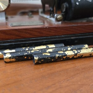 May include: Close-up of black pencils with gold leaf accents. The pencils are lying on a wooden surface, with a vintage machine in the background. The pencils have a hexagonal shape.