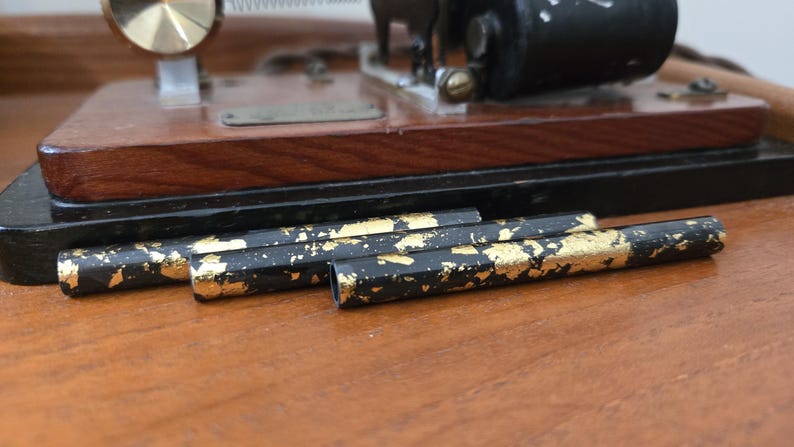 May include: Close-up of black pencils with gold leaf accents. The pencils are lying on a wooden surface, with a vintage machine in the background. The pencils have a hexagonal shape.