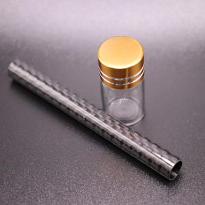 May include: A clear glass vial with a gold-colored lid and a carbon fiber tube. The vial is empty and cylindrical. The tube is black with a woven pattern. Both objects are on a dark surface.
