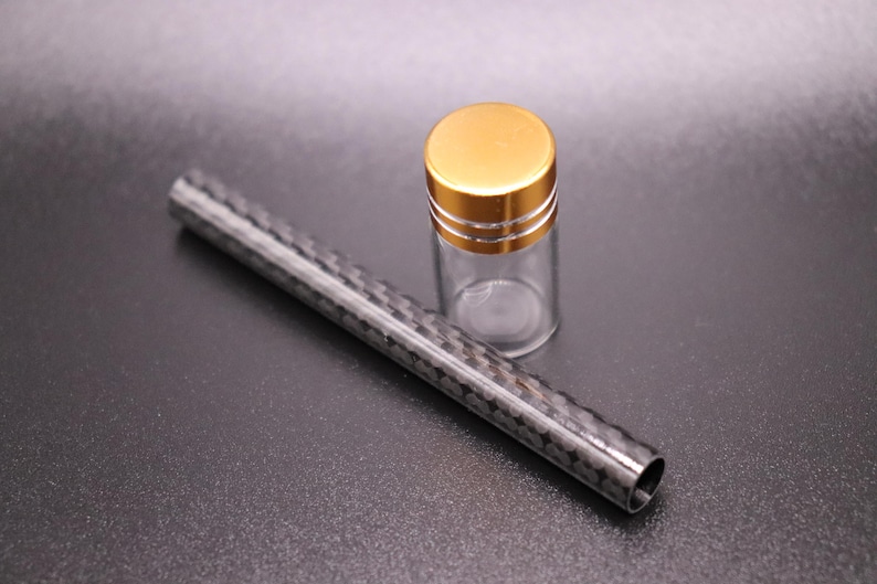 May include: A clear glass vial with a gold-colored lid and a carbon fiber tube. The vial is empty and cylindrical. The tube is black with a woven pattern. Both objects are on a dark surface.