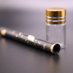 May include: A clear glass vial with a gold-colored lid and a black tube with gold accents. The tube has a hollow opening on one end. The items are on a dark surface.