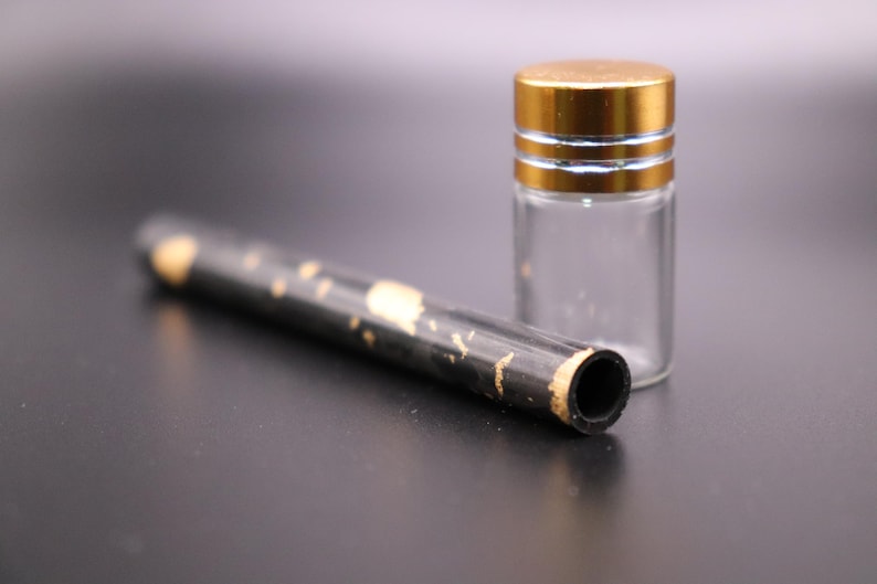 May include: A clear glass vial with a gold-colored lid and a black tube with gold accents. The tube has a hollow opening on one end. The items are on a dark surface.