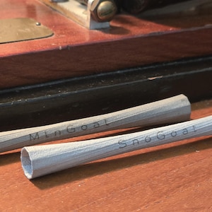 May include: Two grey pencils with the text "Mtn Goat" and "SnoGoat" printed on them, resting on a wooden surface. A vintage machine is visible in the background. The pencils are approximately 15 cm long.
