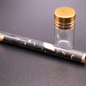 May include: A clear glass vial with a gold-colored lid and a black tube with gold leaf accents. The tube is cylindrical and lies next to the vial. The items are on a dark surface.