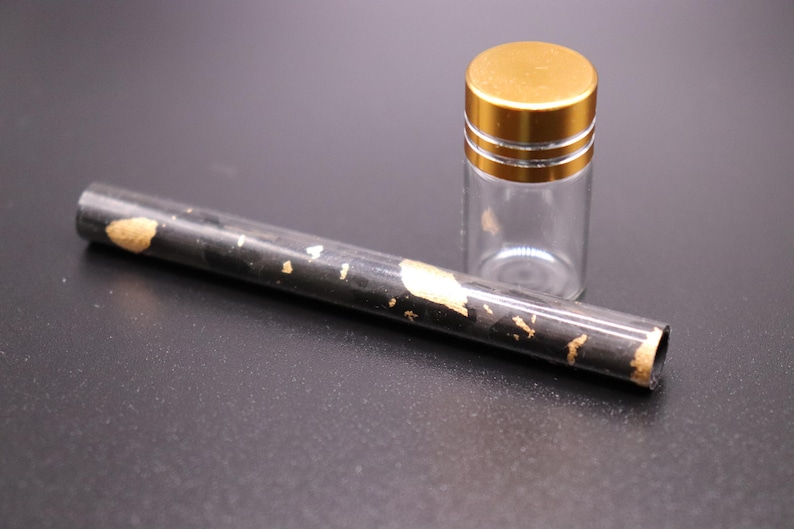May include: A clear glass vial with a gold-colored lid and a black tube with gold leaf accents. The tube is cylindrical and lies next to the vial. The items are on a dark surface.