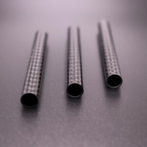 May include: Three carbon fiber tubes with a woven pattern are arranged on a dark surface. The tubes have open ends, revealing a hollow interior. The tubes are cylindrical and appear to be of varying lengths.