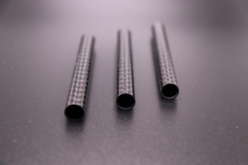 May include: Three carbon fiber tubes with a woven pattern are arranged on a dark surface. The tubes have open ends, revealing a hollow interior. The tubes are cylindrical and appear to be of varying lengths.