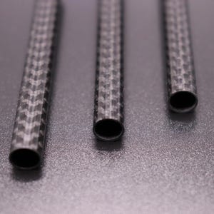 May include: Three carbon fiber tubes with a woven pattern are arranged vertically against a gray background. The tubes have open ends, revealing a dark interior. The textured surface of the tubes reflects light, highlighting the intricate weave.