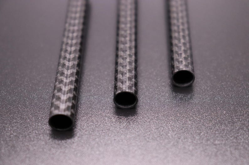 May include: Three carbon fiber tubes with a woven pattern are arranged vertically against a gray background. The tubes have open ends, revealing a dark interior. The textured surface of the tubes reflects light, highlighting the intricate weave.