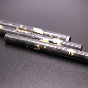 Può includere: Quattro tubi metallici neri con accenti in foglia d'oro sono disposti diagonalmente su una superficie scura. I tubi sono cilindrici e hanno estremità aperte. La foglia d'oro è applicata in modo irregolare, creando un aspetto strutturato.