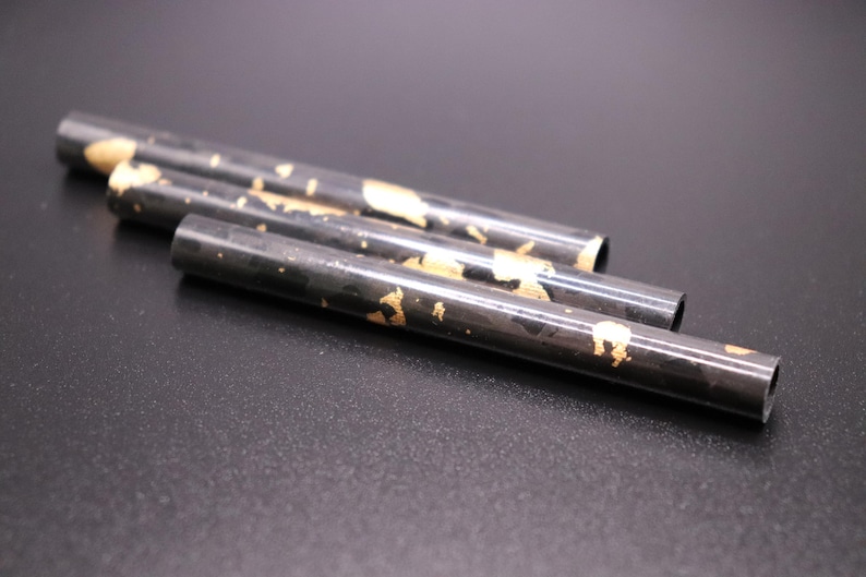 May include: Four black metal tubes with gold leaf accents are arranged diagonally on a dark surface. The tubes have a cylindrical shape and are open at both ends. The gold leaf is irregularly applied.