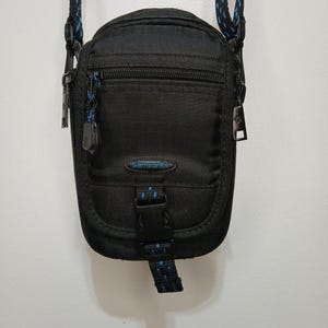 May include: A black vertical crossbody bag featuring a zippered front pocket, a central Samsonite logo patch in blue, and a black plastic buckle closure. The bag includes a black and blue patterned adjustable shoulder strap and metal zipper pulls.