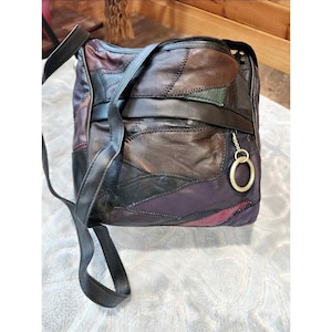 May include: A patchwork leather shoulder bag featuring various shades of brown, black, purple, and dark green leather pieces stitched together. The bag includes a black leather strap, a horizontal black band across the front, and a gold ring charm.