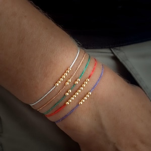 May include: Six colourful string bracelets, each with a row of small, gold-coloured beads. The bracelets are in shades of white, tan, green, red, and purple. The bracelets are worn on a person's wrist.