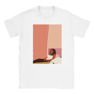 May include: White t-shirt featuring an illustration of a person sitting on a bed. The illustration has a minimalist style with a color palette of pink, beige, and black. The person is wearing glasses and a white top.