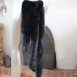 Puede incluir: Un par de colas de piel sintética negra unidas a un arnés. Las colas son largas y esponjosas, y cuelgan del arnés.