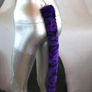 May include: Purple and black striped faux fur tail with a silver clip. The tail is attached to a white elastic band.