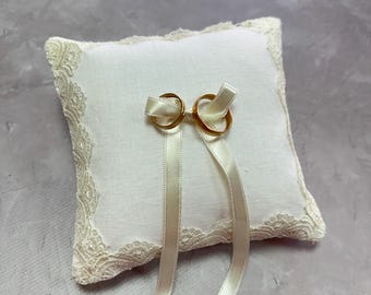 Könnte beinhalten: Ein weißes Kissen mit Spitzenbesatz und einer Schleife aus Satinband. Zwei goldene Eheringe sind an der Schleife befestigt.