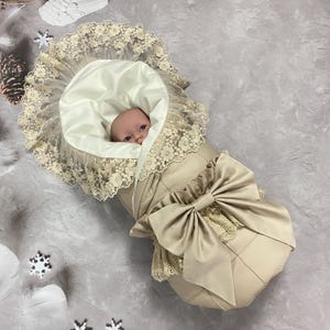 May include: A beige baby sleeping bag with a large bow and lace trim. The sleeping bag has a quilted design and a cream-coloured interior. A baby is nestled inside, with only their face visible.