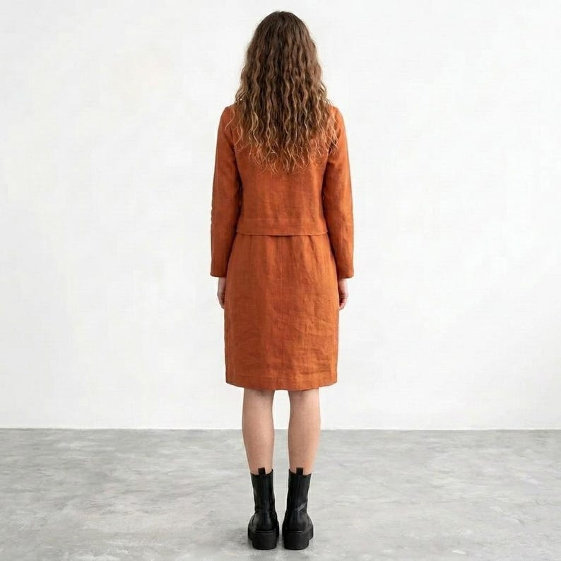 May include: A rust-colored two-piece set consisting of a long-sleeved jacket and a knee-length skirt. The jacket features a straight hemline and long sleeves, while the skirt has a straight cut. The outfit is paired with black ankle boots.