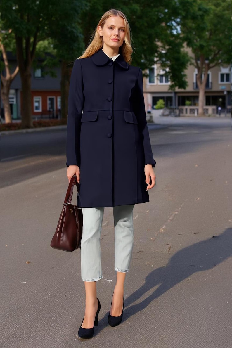 Op de afbeelding: Een marineblauwe jas met een ronde kraag en knoopsluiting. De jas heeft twee voorzakken en wordt gecombineerd met een lichtblauwe cropped broek en zwarte hakken. Een bruine leren handtas wordt vastgehouden.