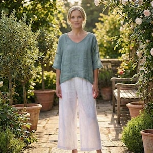 May include: A woman wearing a light green V-neck top and white wide-leg pants. The outfit is made of a lightweight fabric. The background features a garden setting with plants and a wooden bench.