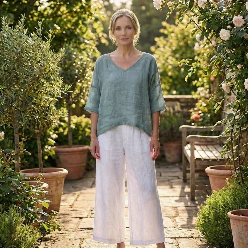 May include: A woman wearing a light green V-neck top and white wide-leg pants. The outfit is made of a lightweight fabric. The background features a garden setting with plants and a wooden bench.