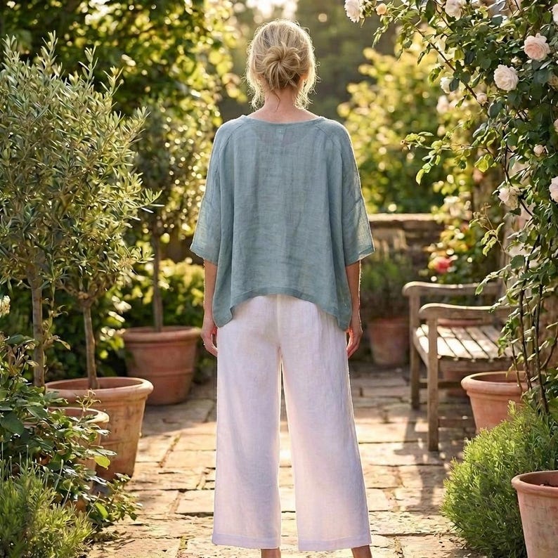May include: A person wearing a light blue linen top and white linen pants. The outfit is loose-fitting and casual. The person is standing in a garden with lush greenery and sunlight.
