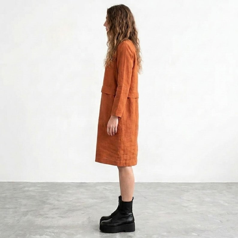 May include: A knee-length, long-sleeved dress in a solid burnt orange color. The garment features a straight silhouette with a horizontal seam detail across the chest and torso area. The dress is paired with black leather platform boots.
